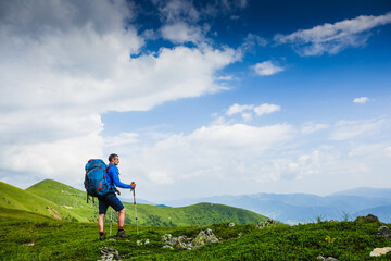 Hiker young man with backpack and trekking poles looking at the mountains in summer outdoor