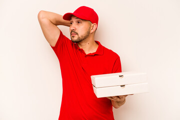 Young delivery man holding pizzas isolated on beige background touching back of head, thinking and making a choice.