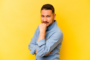 Young hispanic man isolated on yellow background smiling happy and confident, touching chin with hand.