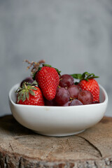 Detalle bol de fresas frescas y  uvas ecológicas  en su punto de maduración con fondo gris y base natural de madera