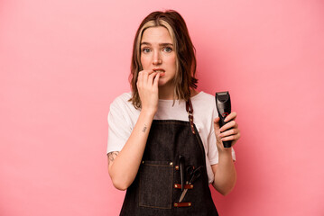 Young hairdresser woman holding scissors isolated on pink background biting fingernails, nervous...