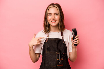 Young hairdresser woman holding scissors isolated on pink background person pointing by hand to a shirt copy space, proud and confident