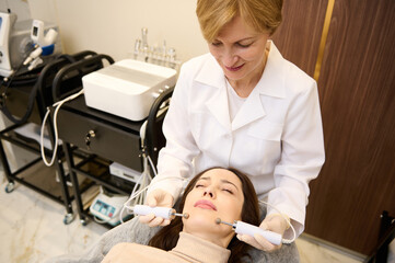 Beautiful middle aged European woman getting stimulating electric facial treatment from beautician during microcurrent therapy in wellness spa clinic