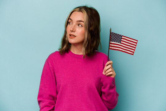 Young caucasian woman holding a American flag isolated on blue background looks aside smiling, cheerful and pleasant.