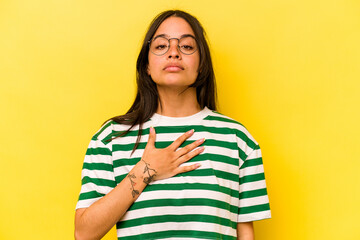 Young hispanic woman isolated on yellow background taking an oath, putting hand on chest.