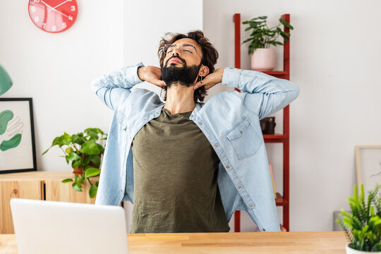 Young hipster freelancer man leaning back in chair doing stretching exercise resting at workplace with hands behind head after successful work. Business and home office concept.