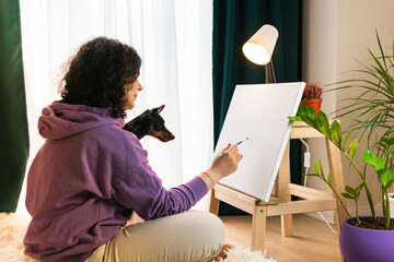 The adult woman painting by numbers indoors, her pet miniature pincher near. People with pets.
