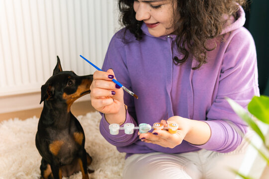The adult woman painting by numbers indoors, her pet miniature pincher near. People with pets.