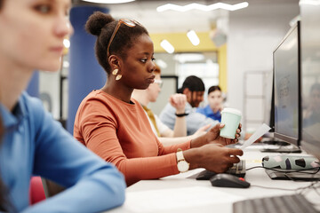 Side view portrait of black young woman using computer in college library while doing research for exams with group of people in row