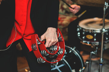Female musician playing tambourine and singing into the microphone, with the musical band performing on stage in the background during jazz rock concert - obrazy, fototapety, plakaty