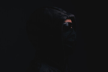 Fototapeta na wymiar Stylish man in dark hoodie with black glasses and mask dark background, hacker, attacker and bandit.
