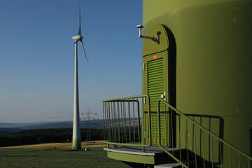 Windkraftanlage vor eine natürlichen Landschaft mit Wäldern und einem Strommast mit einer Tür einer Windkraftanlage im Vordergrund