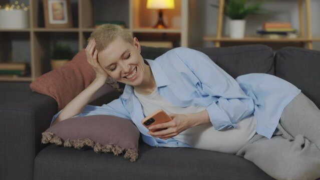 Smiling young woman with big belly lying on comfortable sofa and using modern smartphone. Pregnant blonde laughing while watching some funny videos. Leisure activity of future mother.