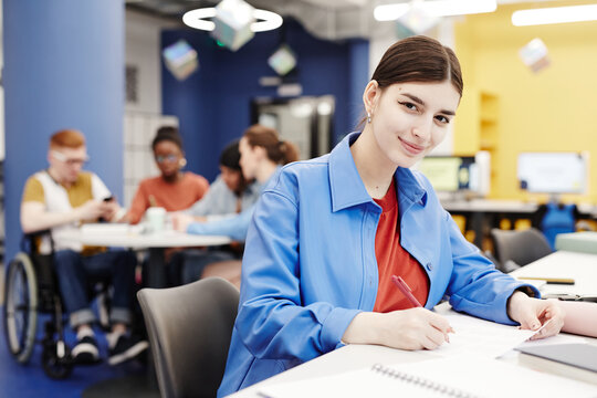 Vibrant portrait of young woman smiling at camera while taking notes in college library
