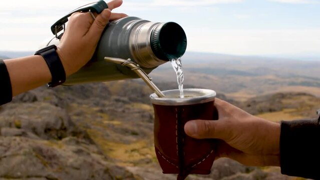 Still horizontal close-up of traditional hot South American caffeine-rich infused beverage mate (yerba mate) .Water poured into steaming mate infusion in red cup, against vegetation and mountain