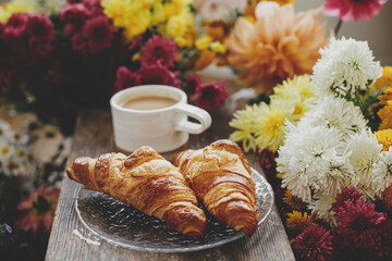 Good morning. Warm cup of coffee and freshly baked croissants on rustic wooden background with beautiful autumn flowers. Stylish mug with cappuccino and croissants on plate, moody image