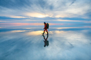 Naklejka na ściany i meble Man traveler with backpack walking on the salt lake at sunset. Sky with clouds are reflected in the mirror water surface. Travel and adventure concept