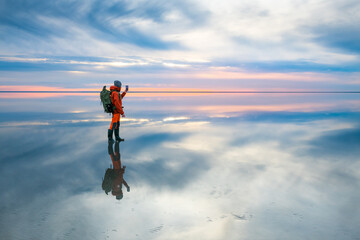 Man traveler with backpack taking photo with smartphone of the beautiful salt lake at sunset. Sky with clouds are reflected in the mirror water surface. Travel and adventure concept