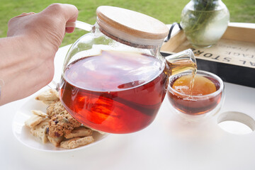 A woman's hand holds a transparent glass teapot with brewed tea and pours tea into a mug. Sunday breakfast in the backyard of the cottage. High quality photo