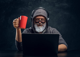 Shot of bearded elderly hacker holding red cup and using laptop against dark background.