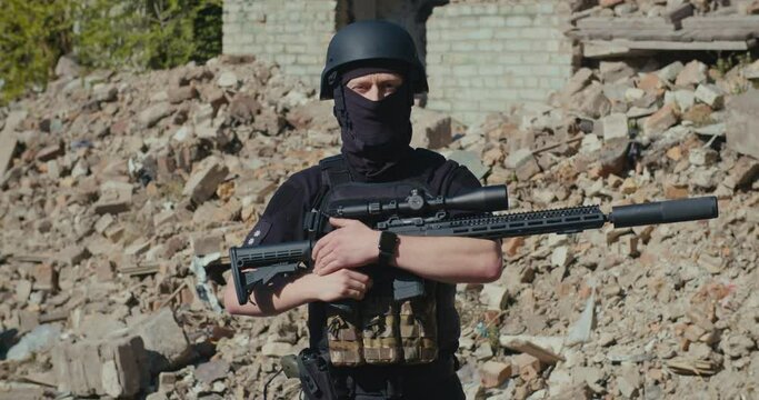 A soldier with a rifle looks around against the background of a destroyed house.