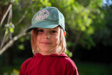 Mały sześcioletni chłopiec w czapce z daszkiem uśmiecha się stojąc na dworze na tle drzew i roślinności - obrazy, fototapety, plakaty