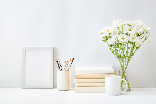 Mockup with a white frame, books, white flowers in a vase, office supplies on a light background. Empty poster frame mockup for presentation design, text, lettering