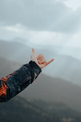 Unrecognizable male hand wearing a coat making the hang loose sign on top of a mountain with peaks in the background