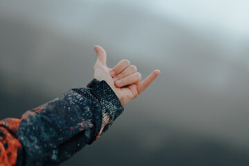 Unrecognizable male hand wearing a coat making the hang loose sign on top of a mountain with peaks in the background