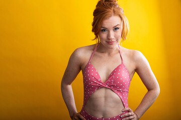 Portrait of woman with red hair and freckles wearing pink polka dot swimsuit. Seamless yellow background