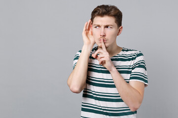 Young curious man 20s wearing blue striped t-shirt try to hear you overhear listening intently says hush be quiet with finger on lips shhh gesture isolated on plain gray background studio portrait