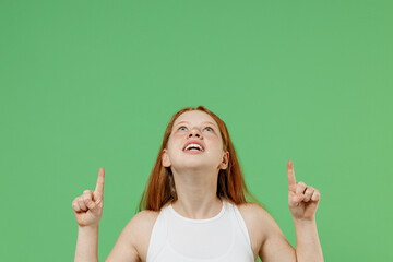 Little redhead kid fun girl 12-13 years old in white tank shirt point index finger overhead on workspace area mock up copy space isolated on plain green background studio Childhood lifestyle concept