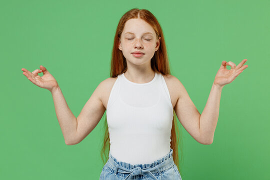 Little redhead kid spiritual girl 12-13 years old in white tank shirt hold spreading hands in yoga om aum gesture relax meditate try to calm down isolated on plain green background studio portrait
