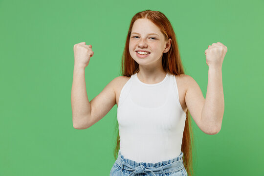 Little redhead kid girl 12-13 years old in white tank shirt doing winner gesture celebrate clenching fists say yes isolated on plain green color background studio portrait Childhood lifestyle concept