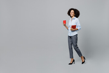 Full body young employee business corporate lawyer woman of African American ethnicity in formal shirt work in office hold takeaway red plastic cup coffee to go hold book isolated on grey background.