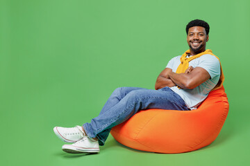 Full body young man of African American ethnicity wear blue t-shirt sit in bag chair hold hands crossed folded look camera isolated on plain green background studio portrait. People lifestyle concept.