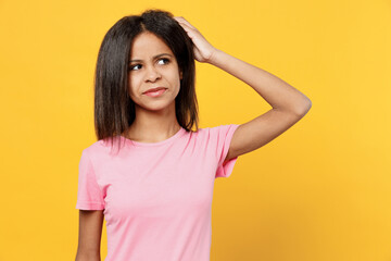 Fototapeta premium Little puzzled sad kid girl of African American ethnicity 12-13 years old in pink t-shirt look aside scratch hold head isolated on plain yellow background studio portrait. Childhood lifestyle concept.