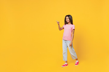 Fototapeta na wymiar Full body happy fun little kid girl of African American ethnicity 12-13 years old wearing pink t-shirt point finger aside on workspace isolated on plain yellow background. Childhood lifestyle concept.