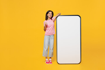 Full body little kid girl of African American ethnicity 12-13 years old in pink t-shirt big huge blank screen mobile cell phone workspace copy space mockup area isolated on plain yellow background