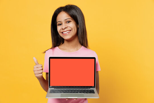 Little kid girl of African American ethnicity 12-13 years old in pink t-shirt hold use work on laptop pc computer with blank screen workspace area show thumb up isolated on plain yellow background