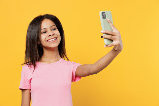 Little kid girl of African American ethnicity 12-13 years old in pink t-shirt doing selfie shot on mobile cell phone isolated on plain yellow background studio portrait. Childhood lifestyle concept.