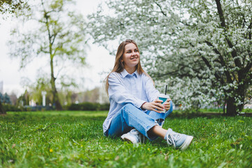 Young hipster woman in denim jacket is resting on green lawn in park, drinking coffee, relaxing outdoors. Lifestyle, coffee break. Selective focus on female hands holding disposable cup of coffee.
