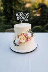 A beautiful two-tier wedding cake with the word Mr and Mrs, decorated with flowers, stands on a table with a white tablecloth. Food photography.