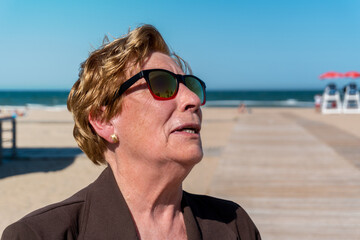 Portrait of an older woman, with a smiling and happy face, wearing fun and colorful sunglasses, on a sunny day at the beach.