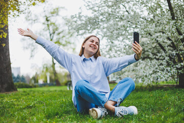 Fototapeta premium Happy woman walking using smart phone in a city street. Business woman with sunglasses uses mobile phone outdoors under blooming tree, girl typing message, chatting, scrolling web page, look at camera