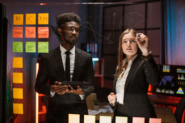Team of young smart multiracial male and female office workers, standing near transparent glass board with colorful steakers and drawing graphs and charts for their new business project