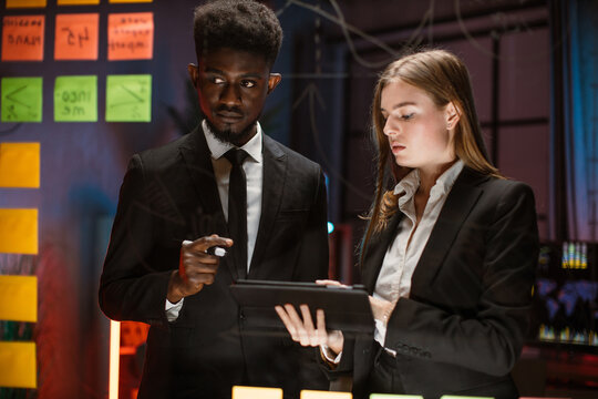 Night work at office, project deadline. Male and female multiethnic creative colleagues, using digital tablet and discuss project with sticky notes on a glass wall.