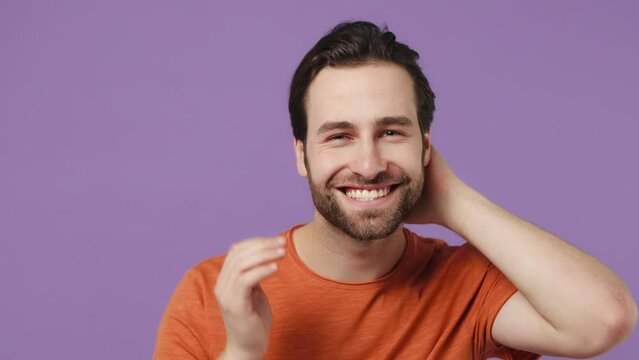 Close up smiling happy young brunet bearded man 20s wears red t-shirt look camera like in mirror beautify himself touch face with hand isolated on plain pastel light purple background studio portrait