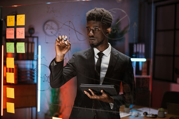 Handsome smart African business man, writing ideas on glass board, working in the night at office...