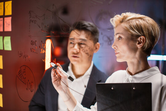 Close up view through glass wall of asian and caucasian colleagues working together in office, searching solution of problem, writing on board with sticky notes, analyzing financial charts